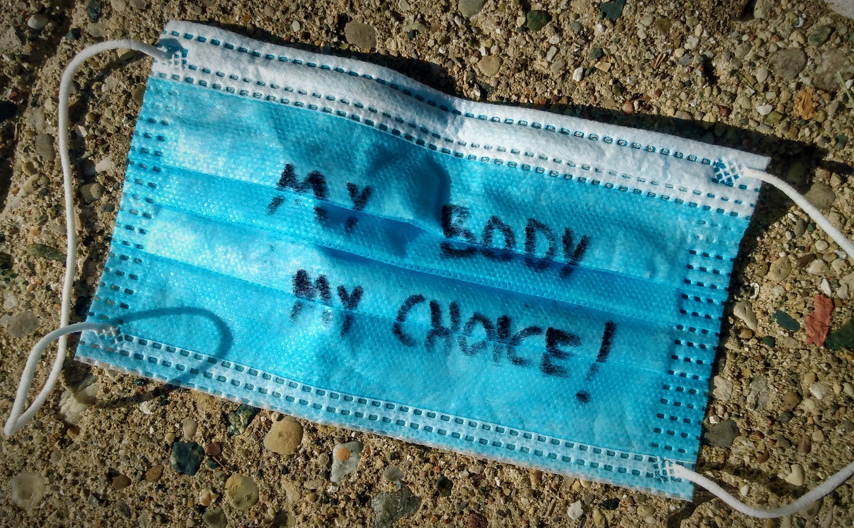 A discarded medical face mask laying on the pavement. It says, "My Body, My Choice!"