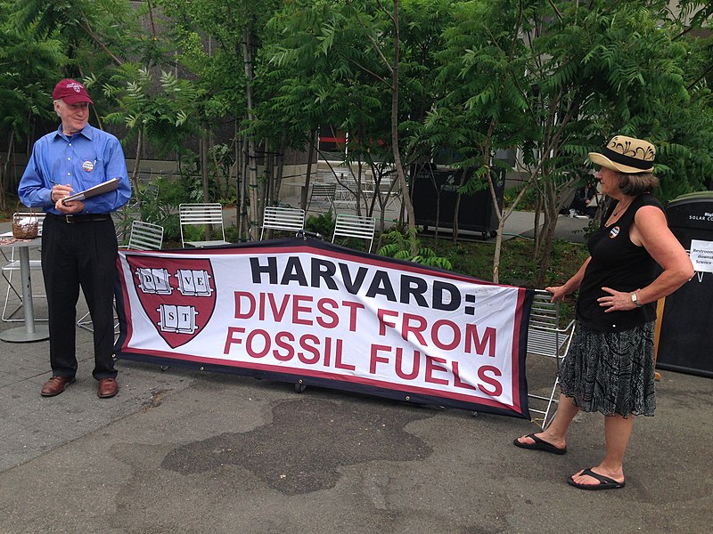 A man and woman stand on either end of a sign that says "Harvard: Divest From Fossil Fuels."