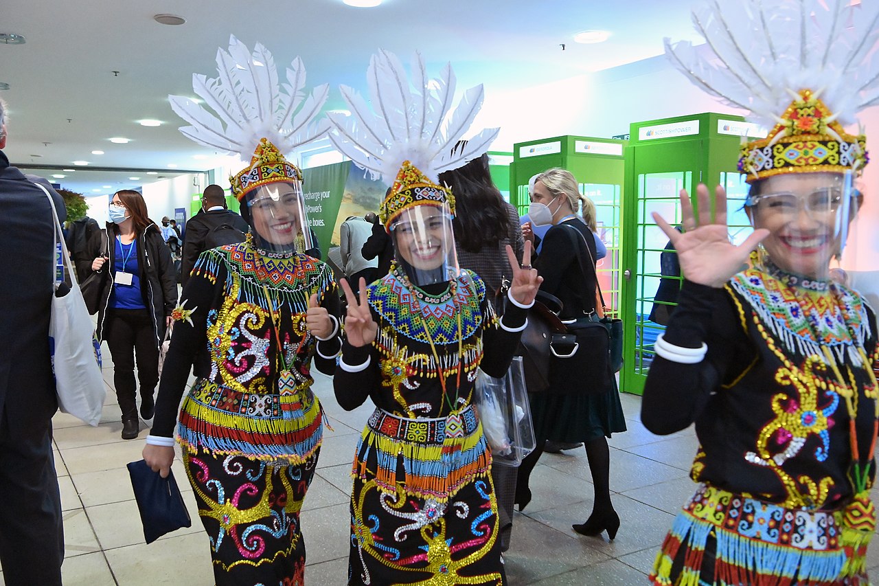 A trio of women in Indigenous regalia and face shields waves and flashes the Victory sign in a hallway at the COP26 conference.
