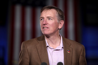 A middle-aged man in a brown jacket in front of a draped American flag.