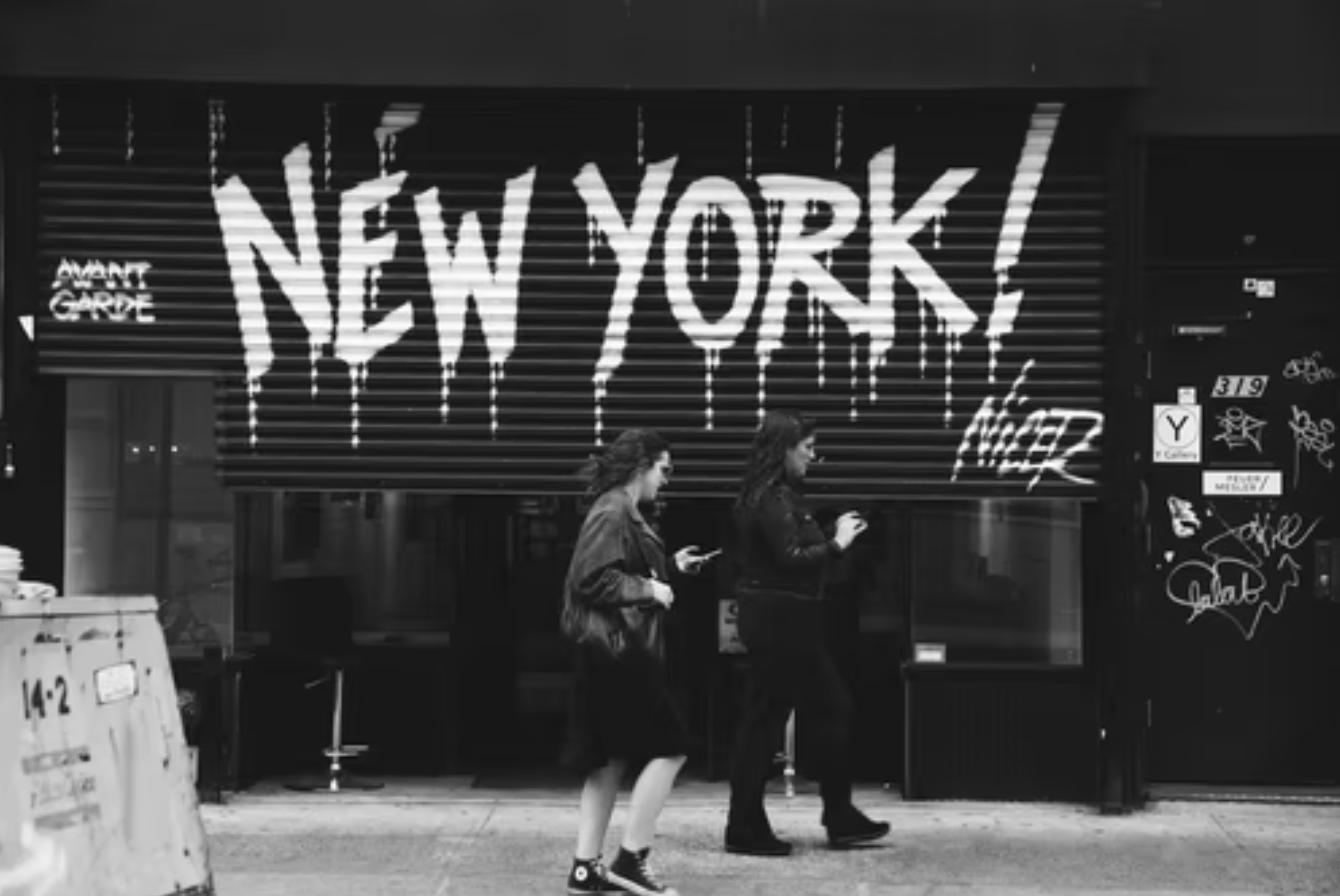 Grey scale of two people walking past door with New York spray painted on it; image by Ian Dooley, via Unsplash.com.