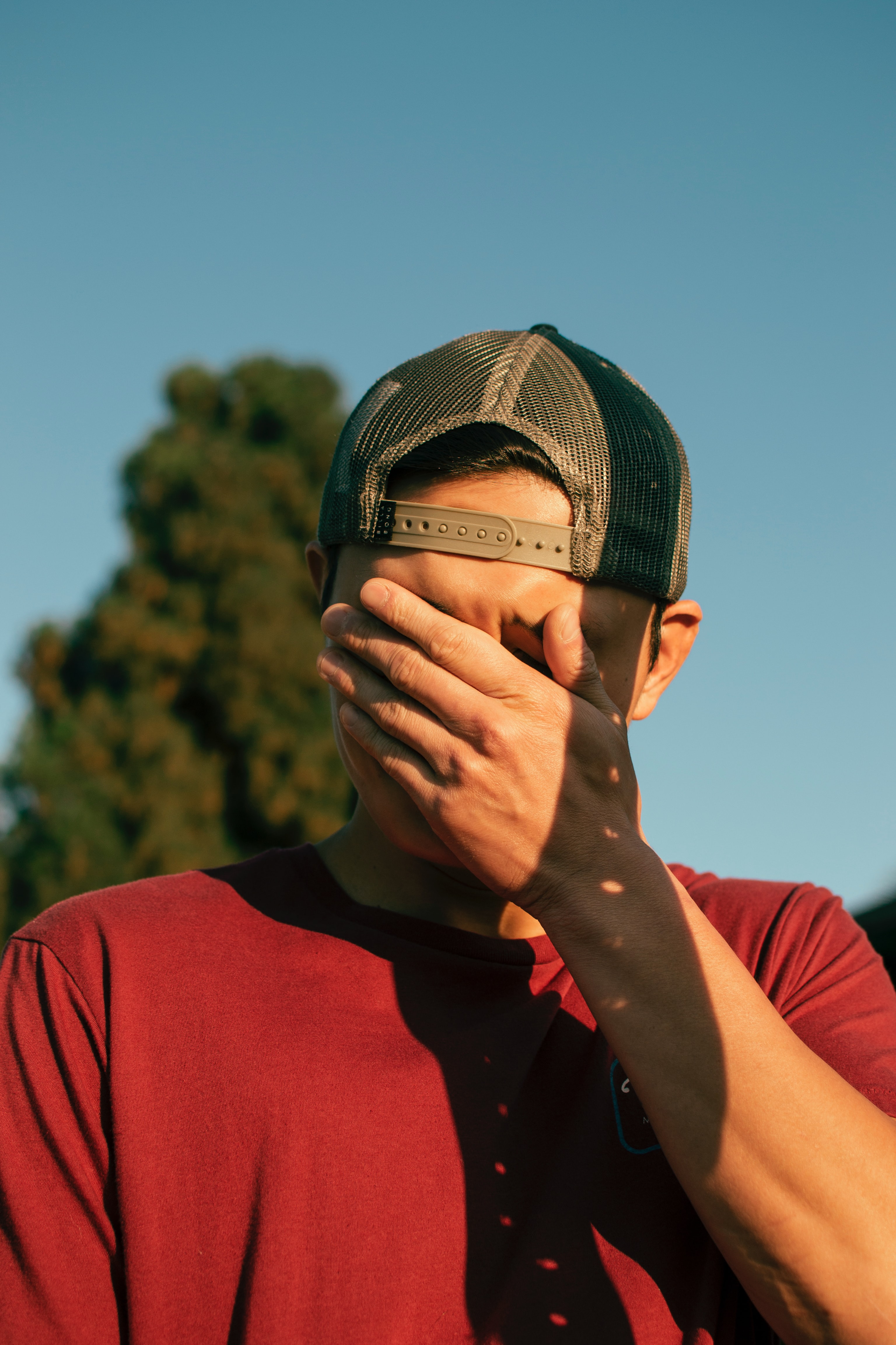 Man in backwards ball cap and maroon shirt with hand over his face; image by Chandra Oh, via Unsplash.com.