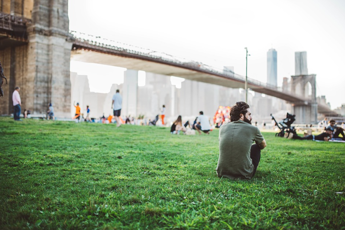 Man sitting on grass in a park by a bridge; image by StockSnap, via Pixabay.com.