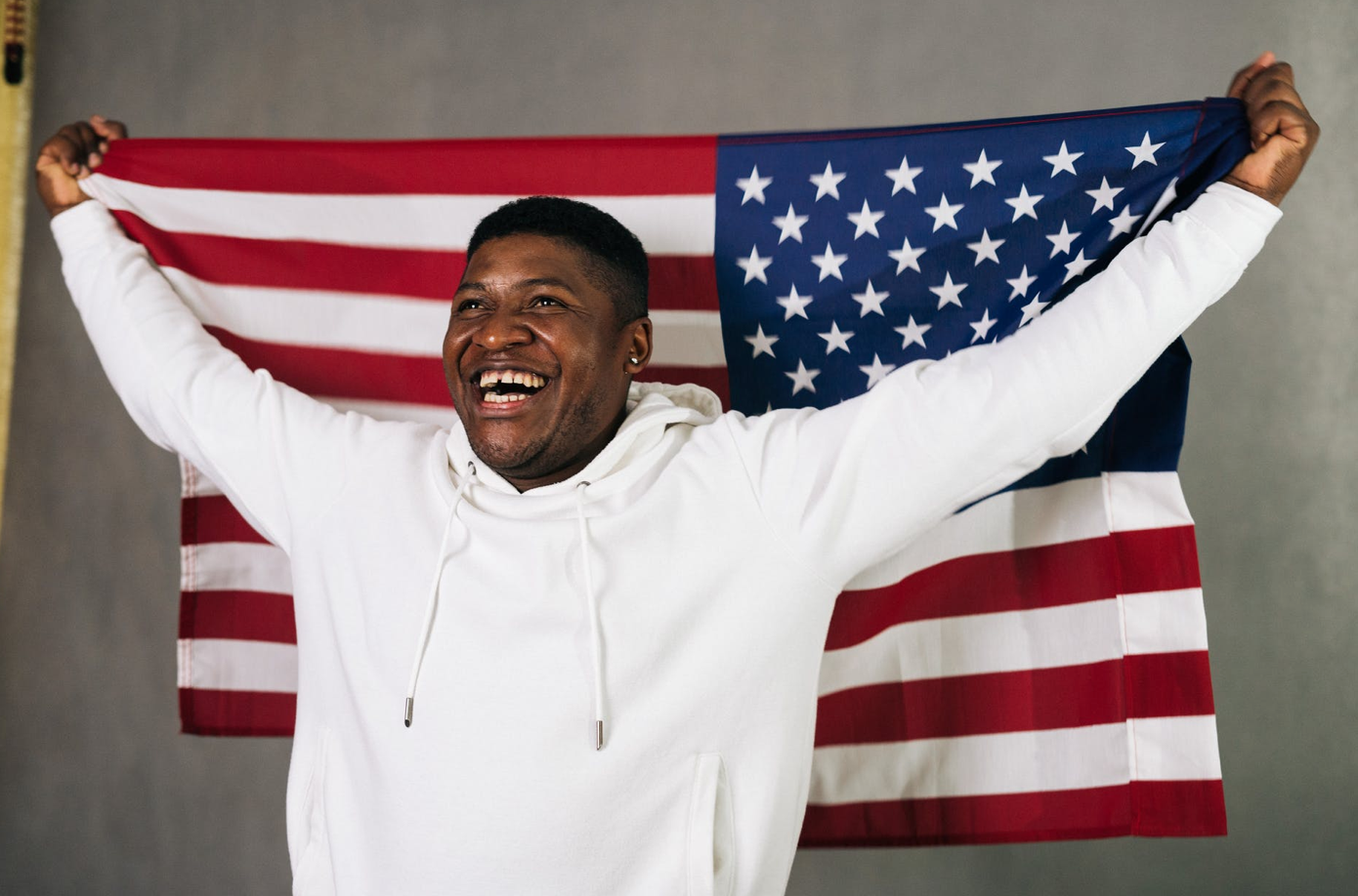 Smiling man in white hoodie holding American flag; image by Tima Miroshnichenko, via Unsplash.com.