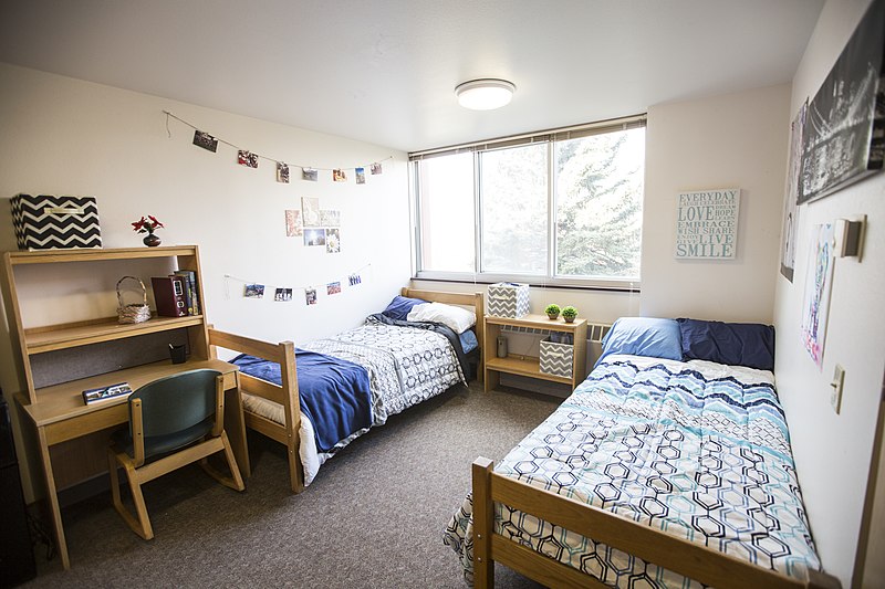 A dorm style room with two beds, a desk, a large window, and posters.