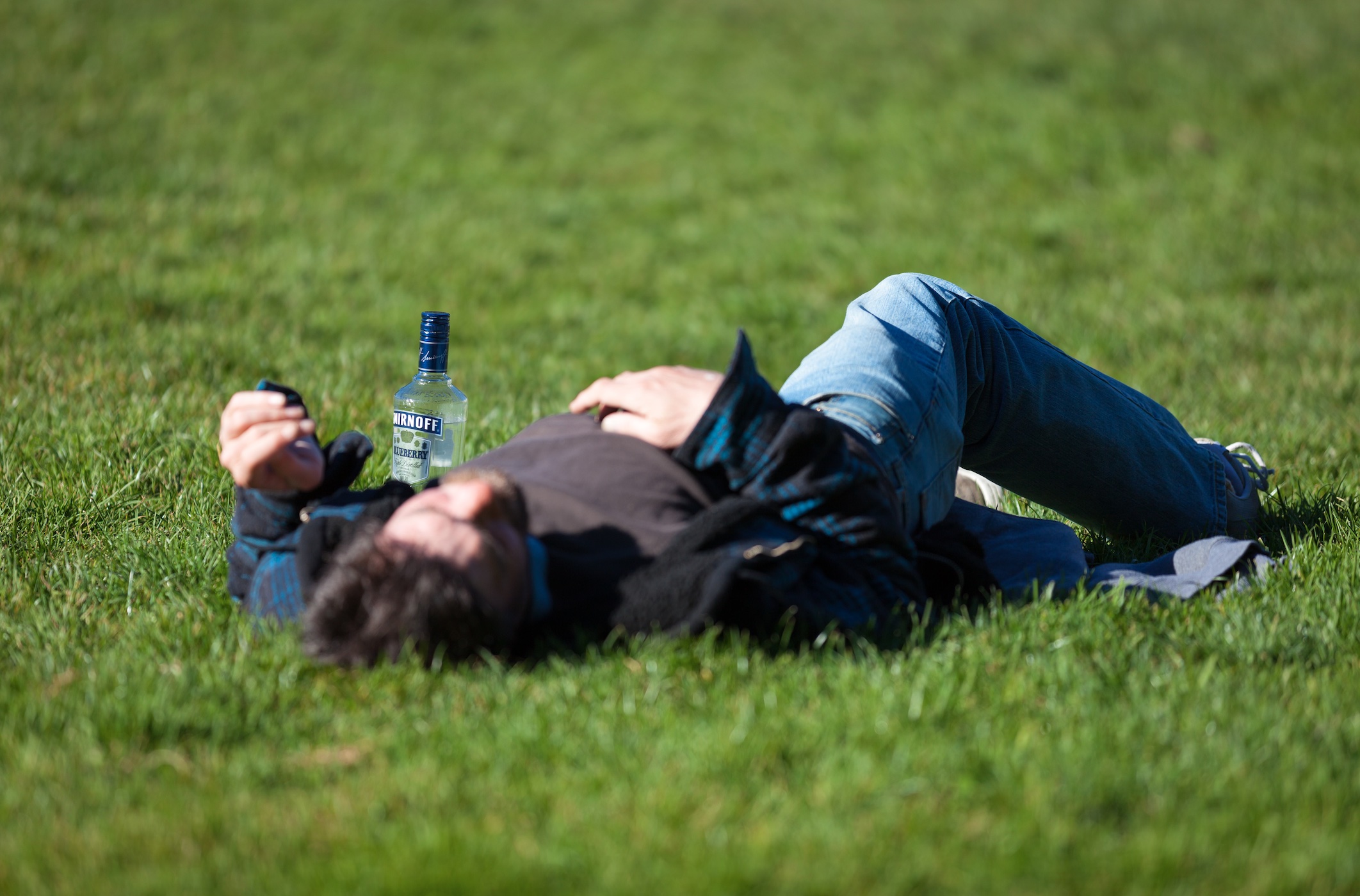 Man passed out on lawn in daylight, empty bottle at his side; image by Thom Masat, via Unsplash.com.