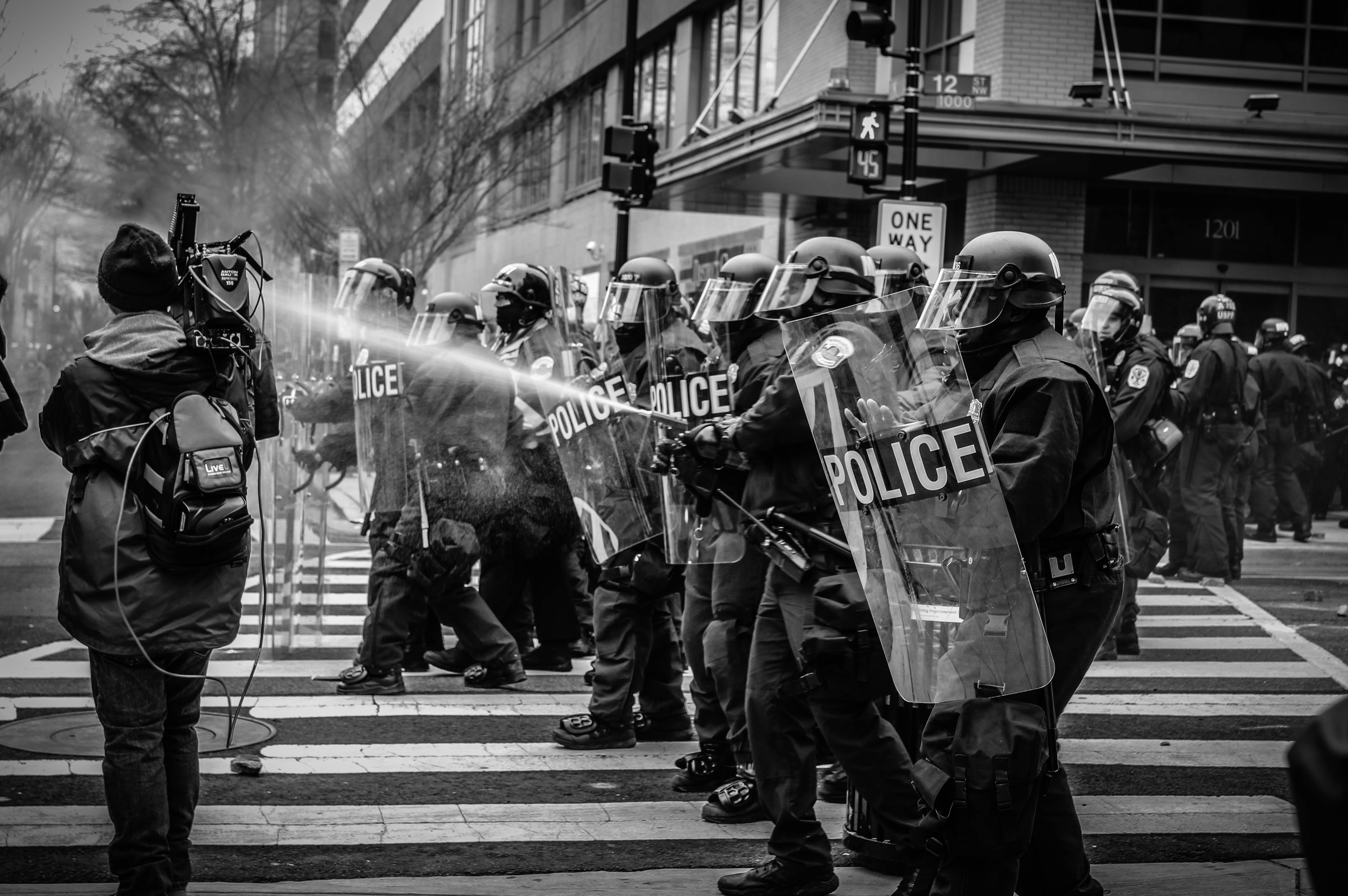 Police in riot gear, one spraying what looks like pepper spray; image by Spenser H, via Unsplash.com.