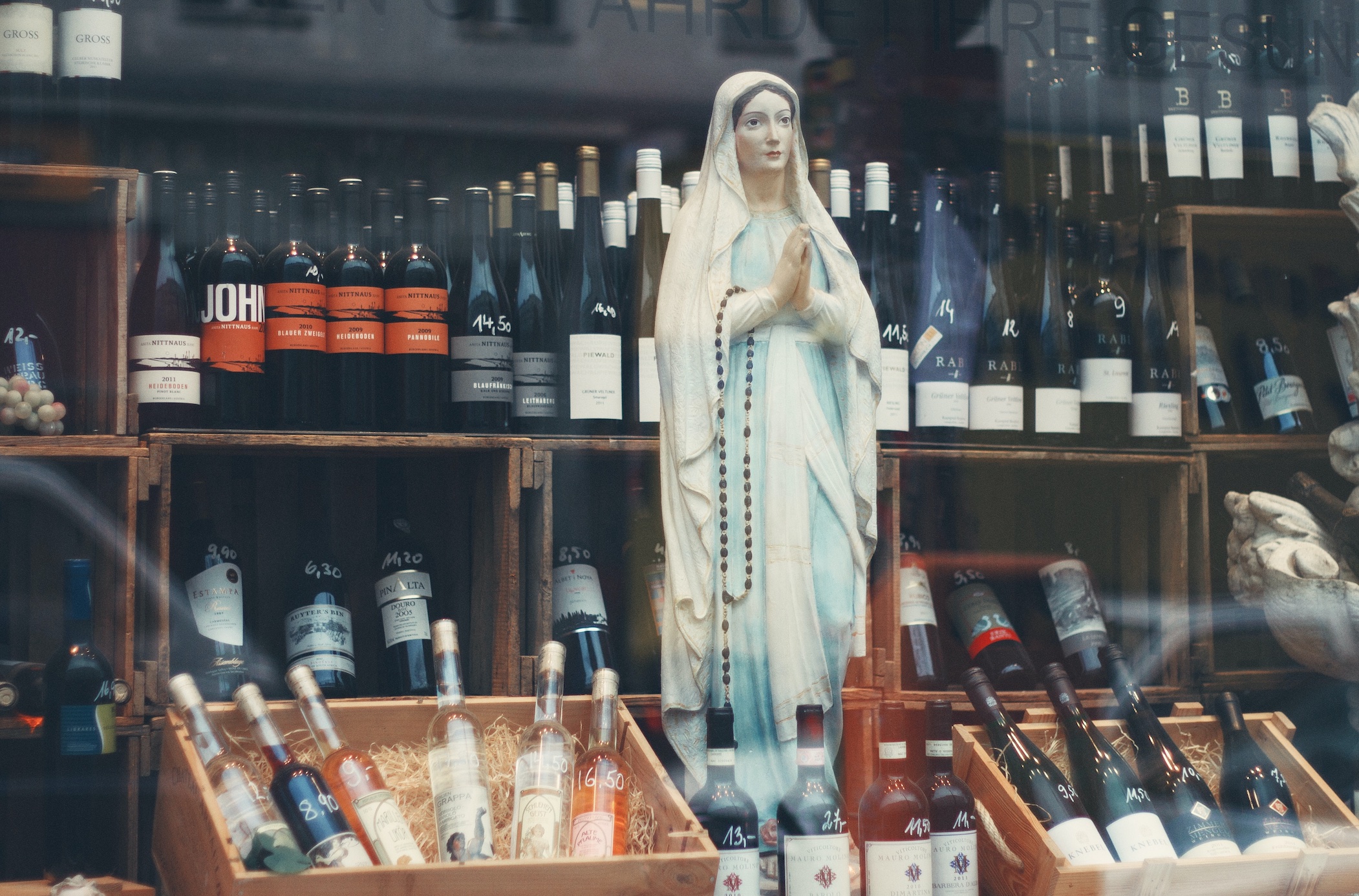 Statue of the Virgin Mary among wine bottles at store; image by Alina Sofia, via Unsplash.com.