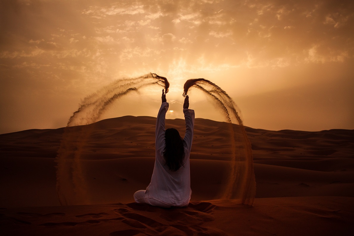 A female figure sits on a sand dune, the wind blowing arcs of sand through her fingers.