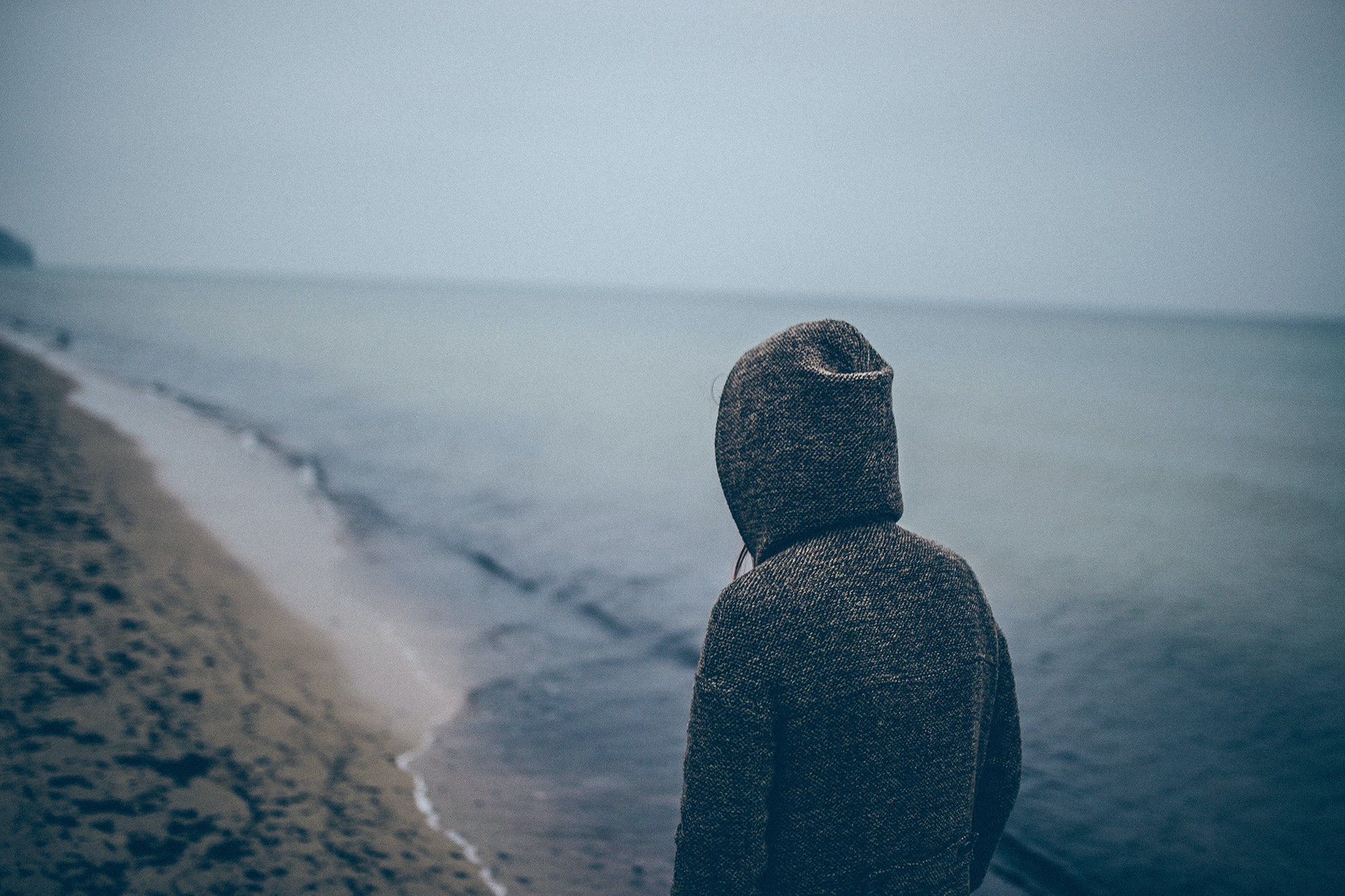 A hooded figure seen from behind walks along a grey and lonely shoreline.