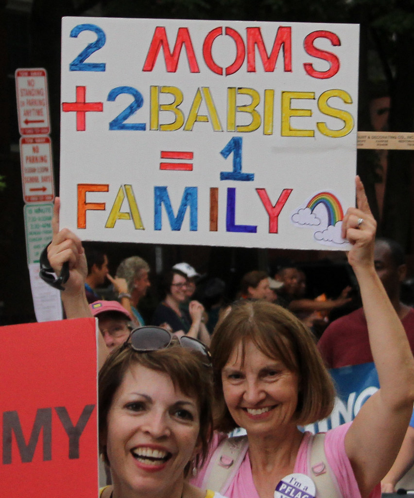 Two woman at a gathering carry a sign that says, "2 Moms + 2 Babies = 1 Family".