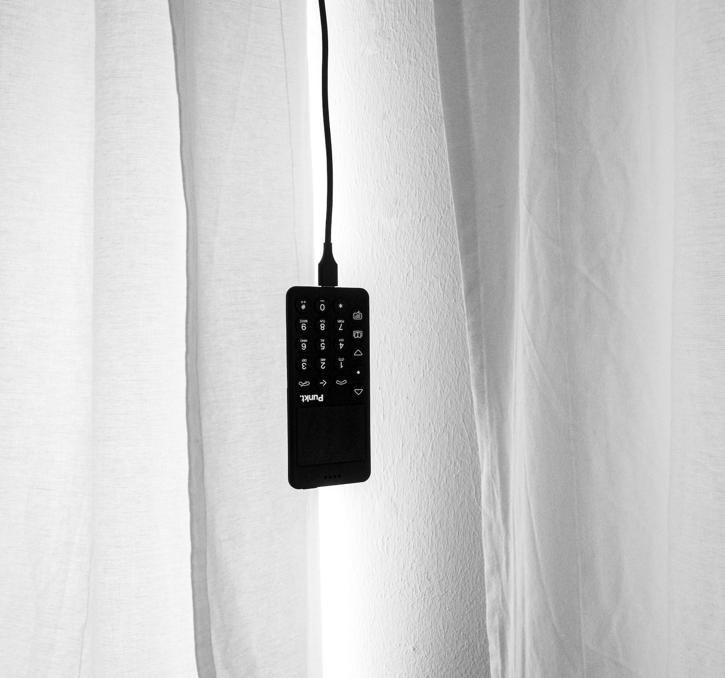 Black cellphone hanging by charging cord aganist white background; image by Thanos Pal, via Unsplash.com.