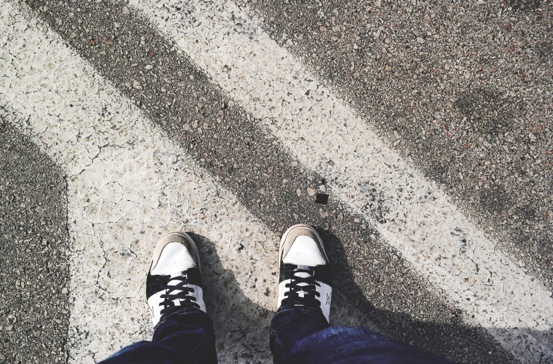 Man in black and white sneakers standing at a crosswalk; image by revac film's&photography, via Unsplash.com.