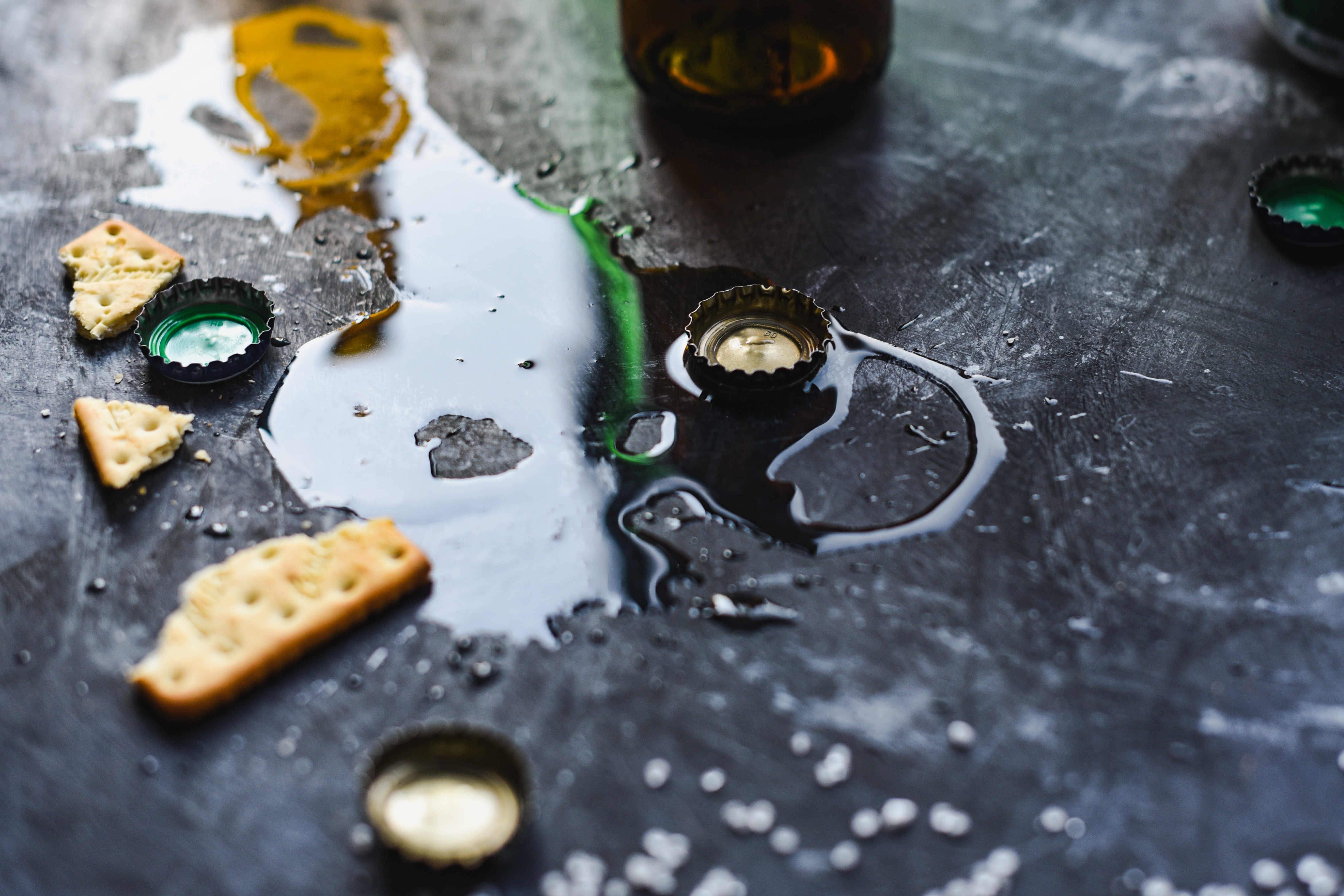 Spilled beer on a black background with beer bottle caps and crakers; image by Anshu A, via Unsplash.com