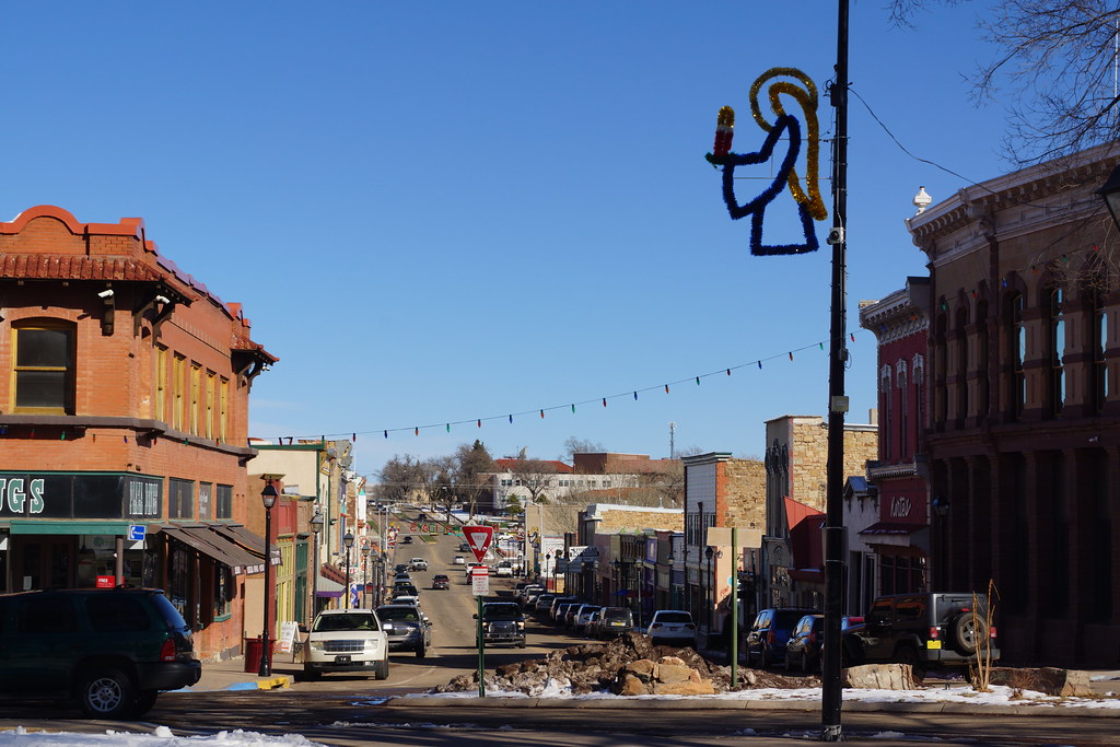 A main street runs through a small town's downtown area, with one-and two-story buildings, storefronts, and traffic, overseen by an angelic holiday decoration.