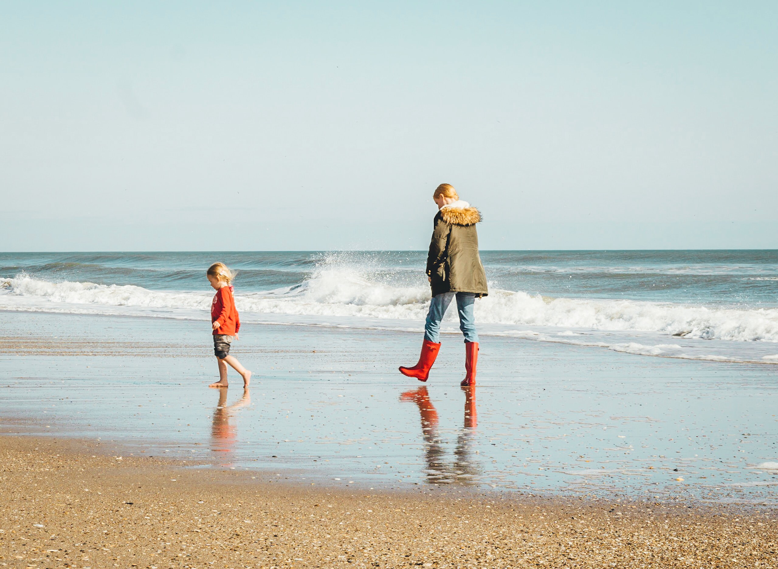 Mom in parka and boots walking on beach with barefoot toddler in shorts; image by Brian Gordillo, via Unsplash.com.