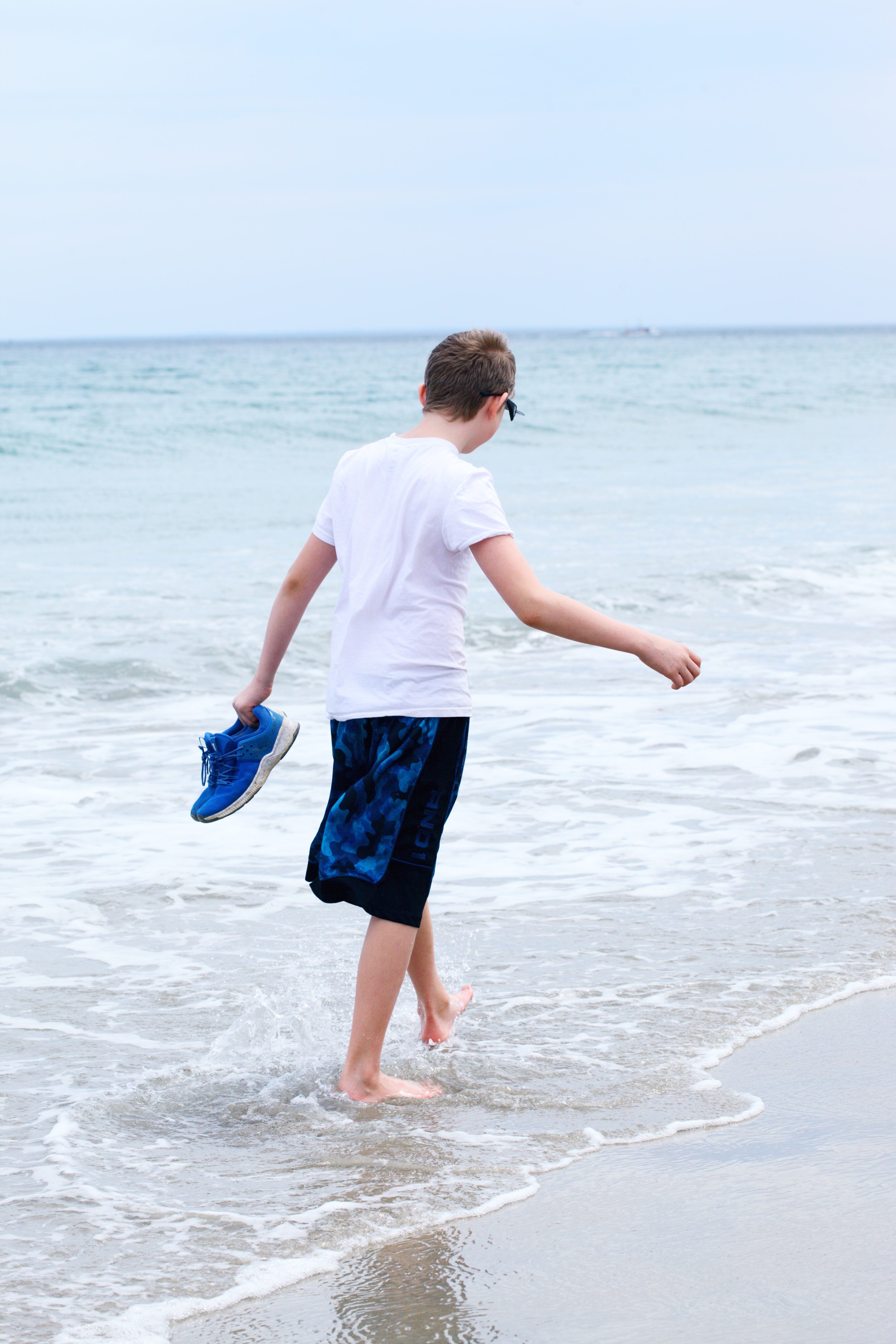 Teen boy in shorts and t-shirt walking in surf carrying blue tennis shoes; image by Alexander Grey, via Unsplash.com.