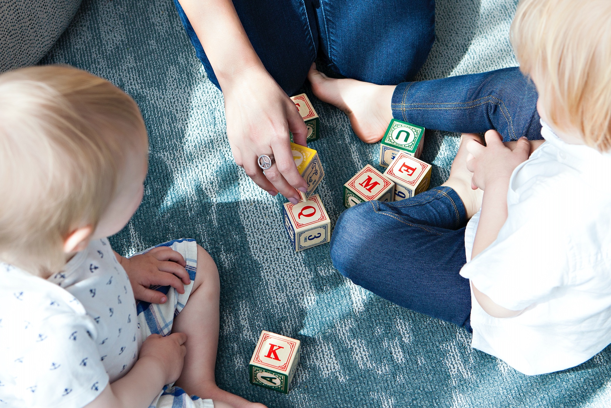 Woman and two little children playing with alphabet blocks on the floor; image by Marisa Howenstine, via Unsplash.com.