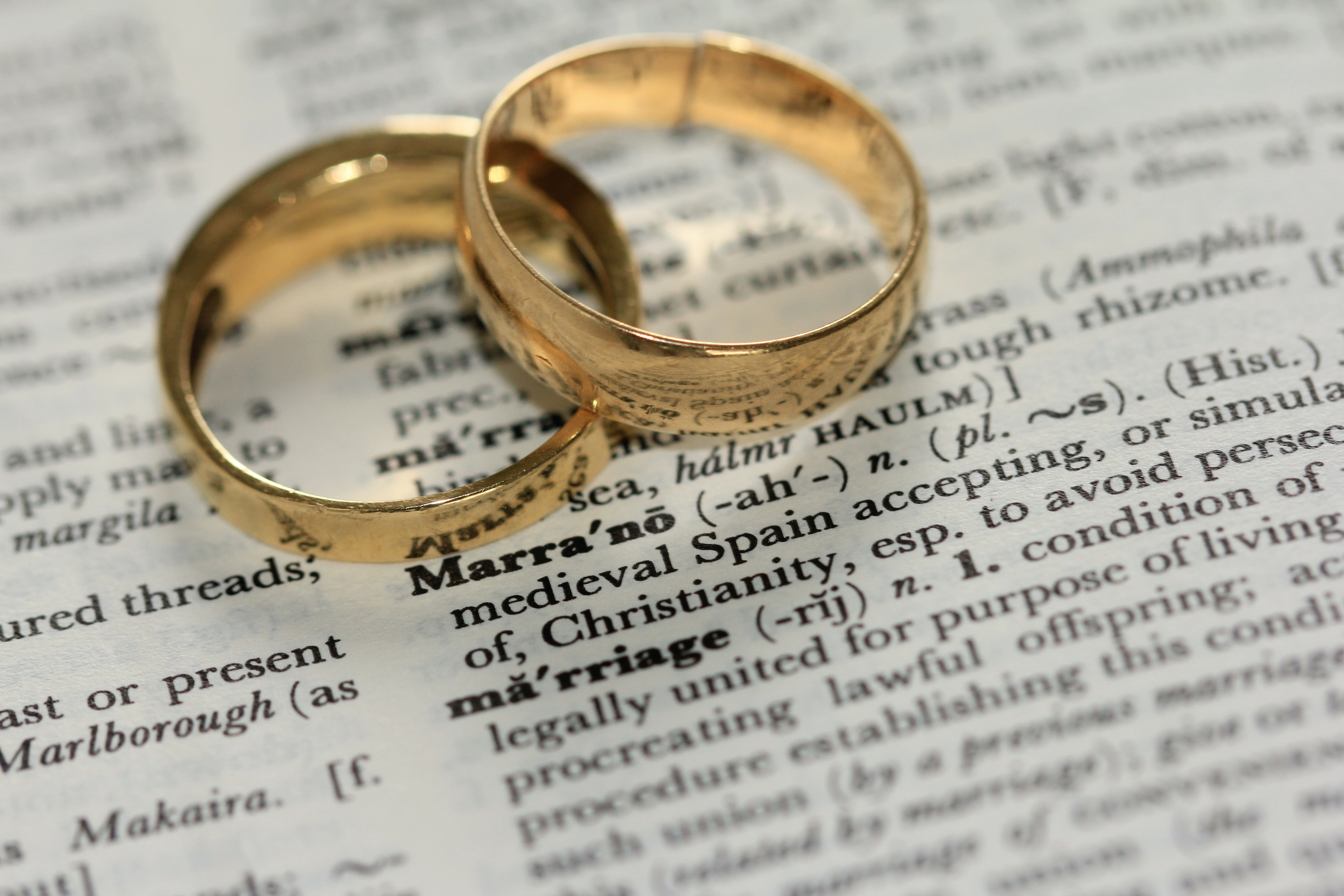 Two gold wedding bands atop a dictionary page defining marriage; image by Sandy Millar, via Unsplash.com.