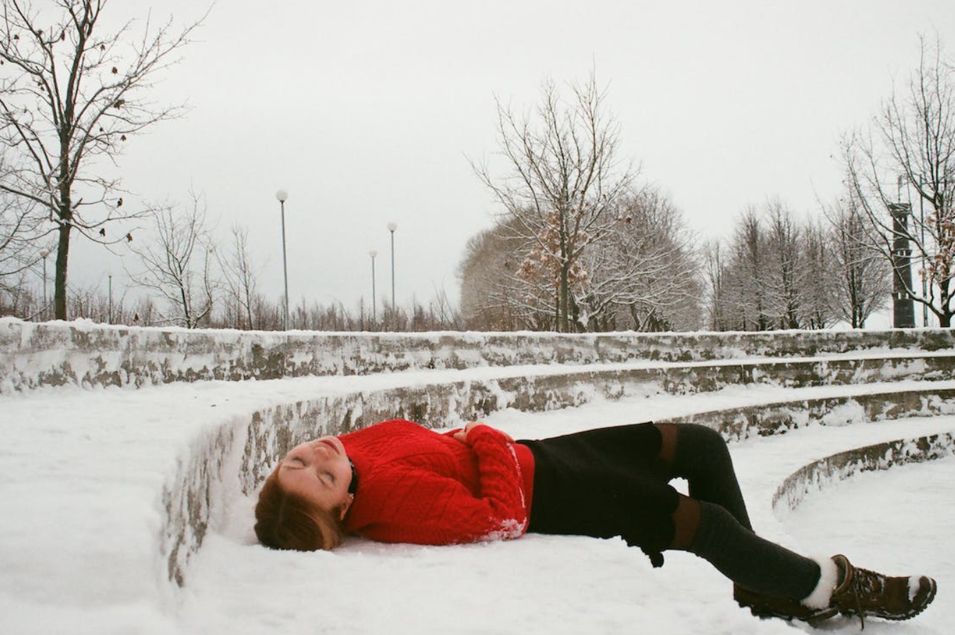 Woman in red coat and black pants laying on snow-covered steps; image by Darya Sannikova, via Pexels.com.