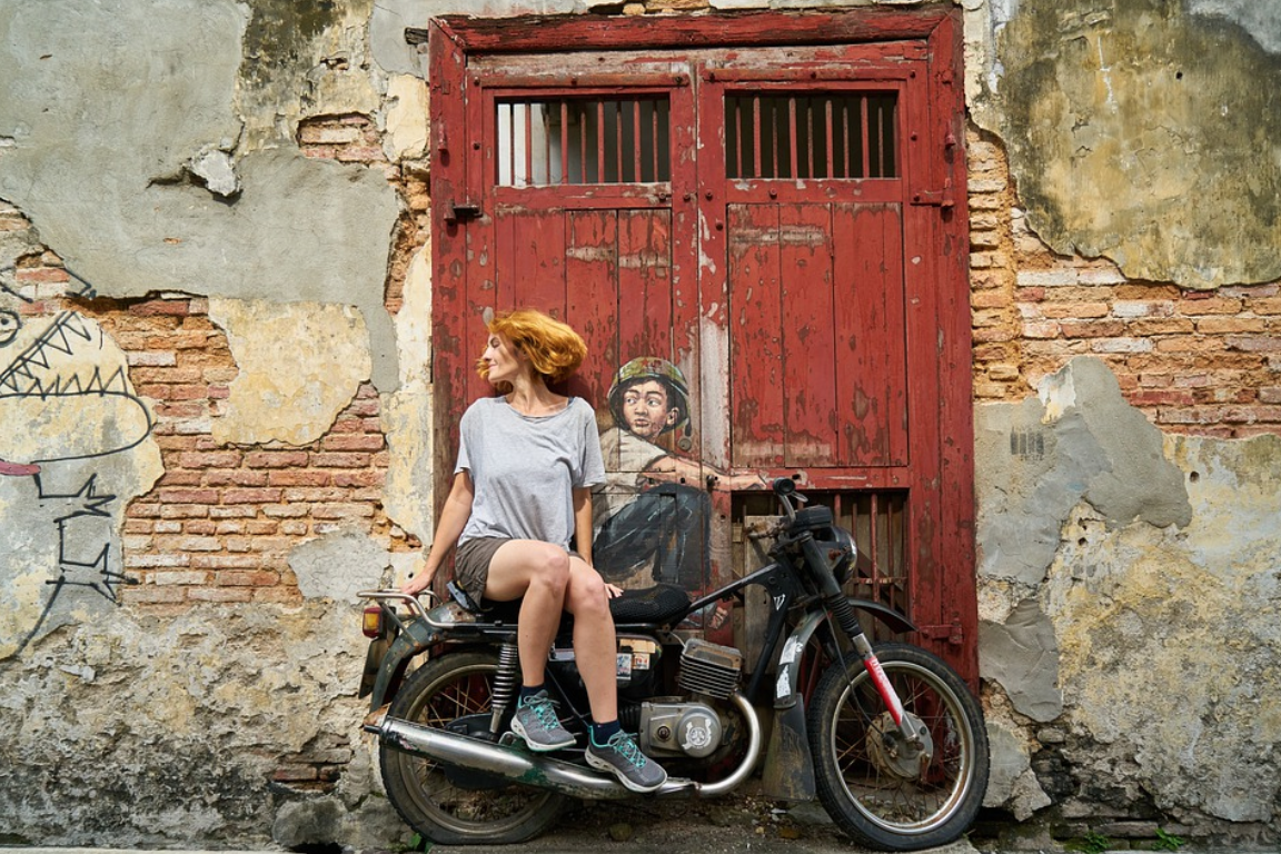 Woman sitting on motorcycle next to mural of motorcycle rider; image by Engin Akyurt, via Pixabay.com.
