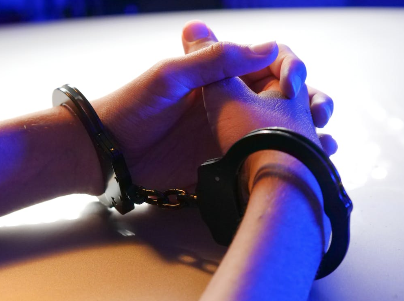 Person in handcuffs resting hands on table; image by Kindel Media, via Pexels.com.