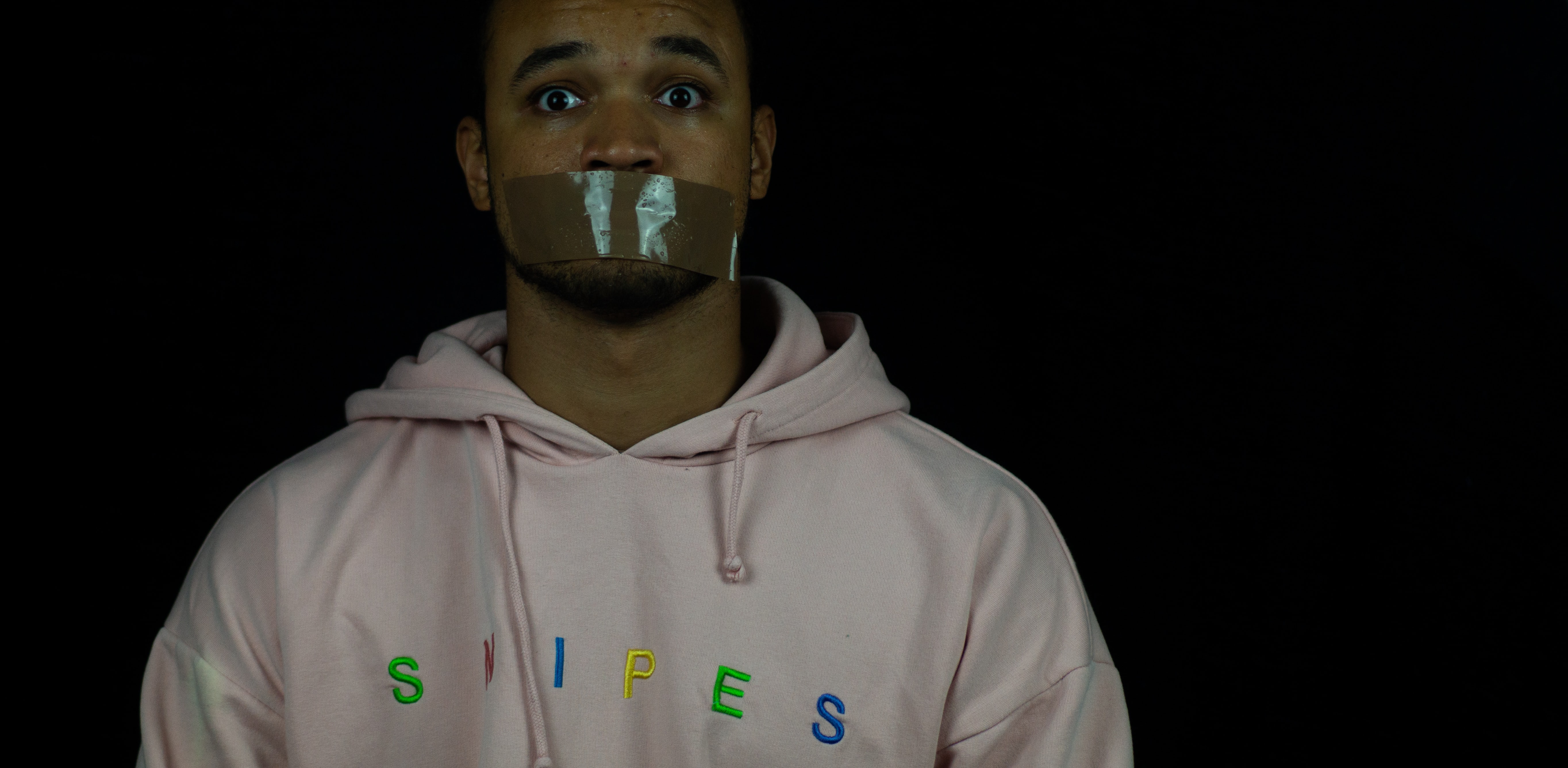 Man in white hoodie with mouth taped shut; image by Khadim Fall, via Unsplash.com.