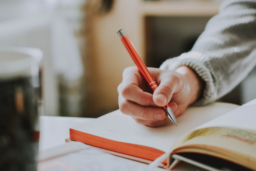 Person holding red pen and writing in book; image by LilArtsy, via Unsplash.com.