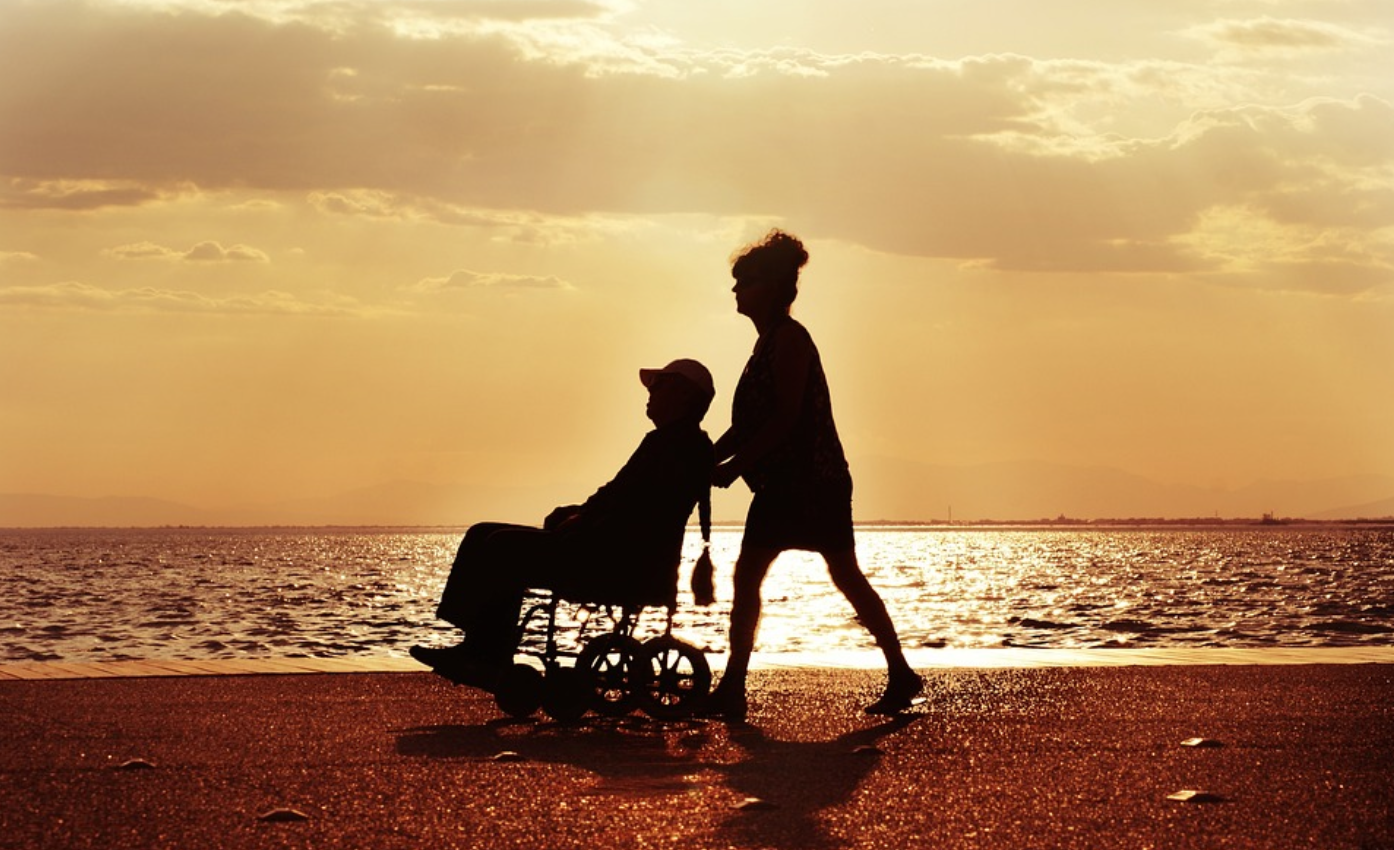 Woman pushing man in wheelchair on beach; image by AdamTepl, via Pixabay.com.