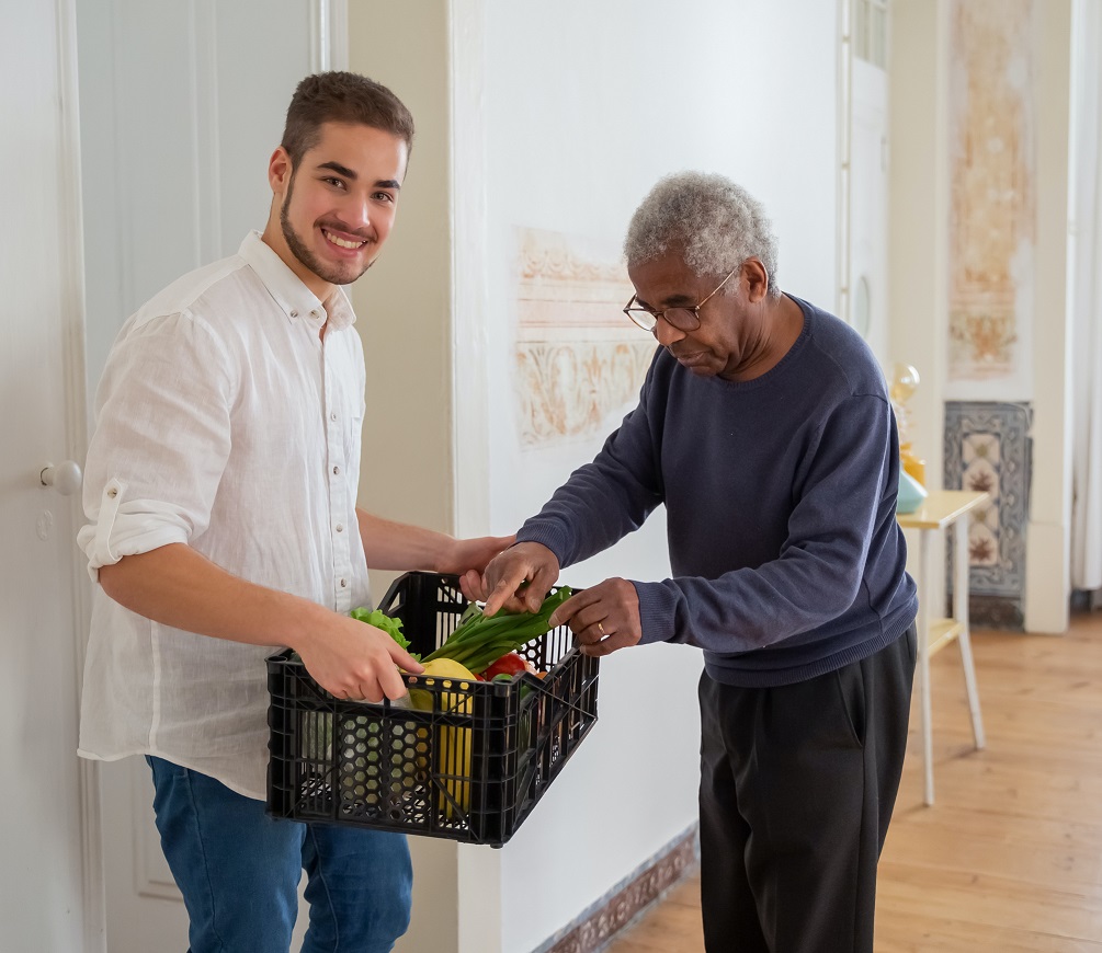 A young man holds a crate of vegetables while an elderly man examines them.