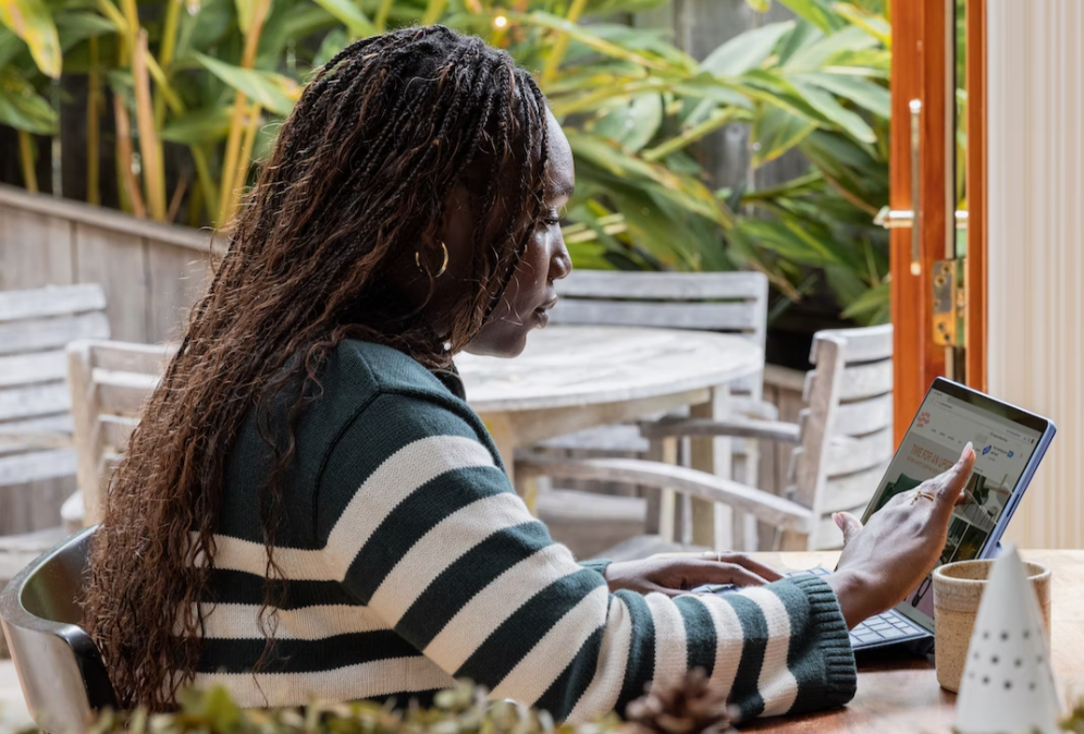 Woman with touchscreen laptop sitting at outdoor table; image by Microsoft Edge, via Unsplash.com.