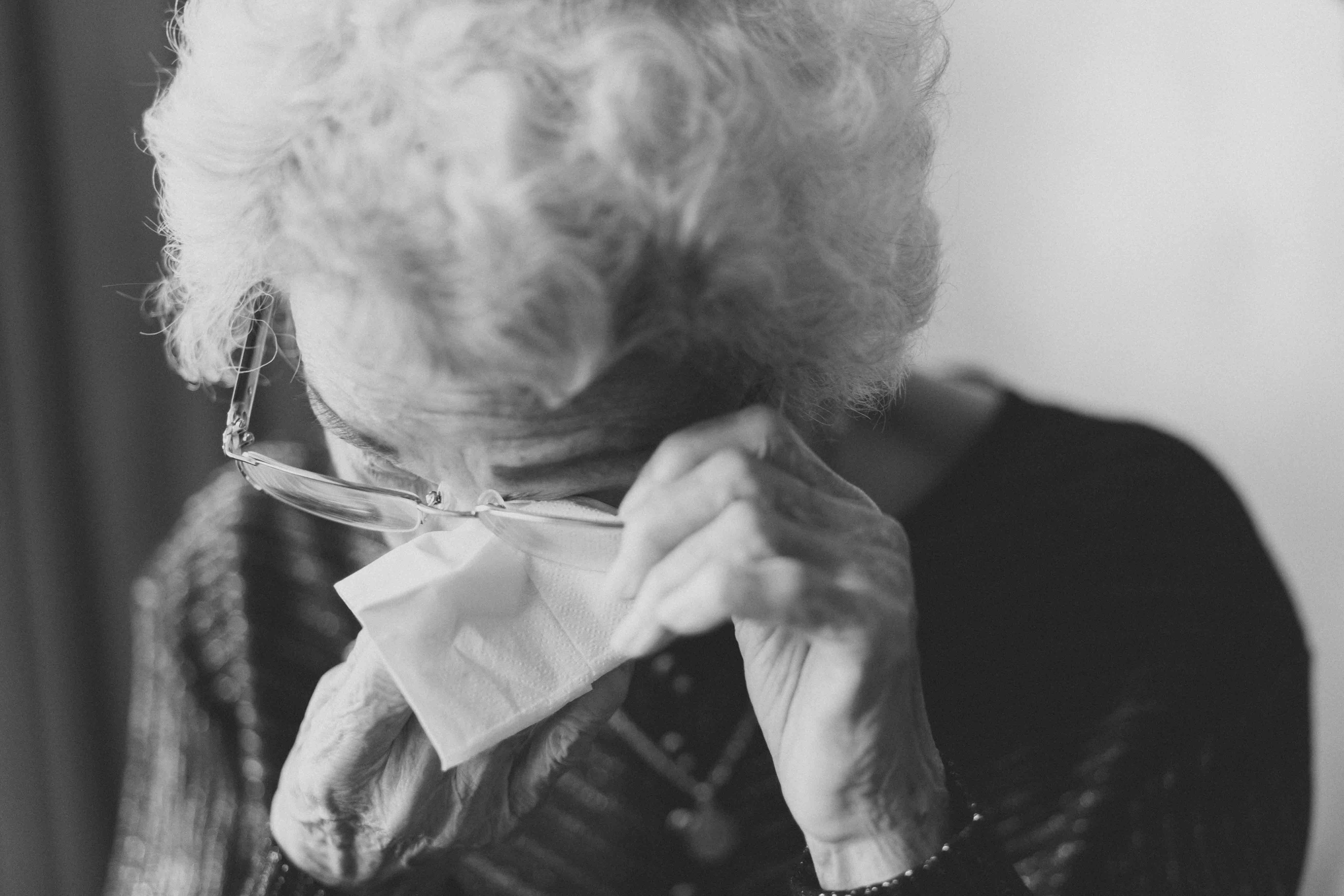 Elderly woman in glasses wiping eye with tissue; image by Jeremy Wong, via Unsplash.com.