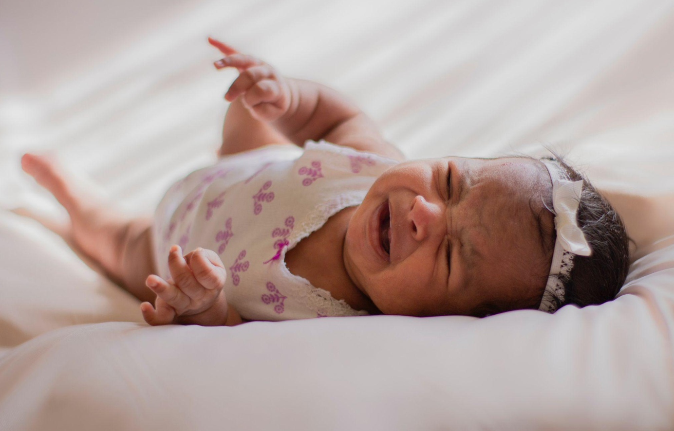 Baby in a onesie on a bed, crying; image by Laura Garcia, via Pexels.com.