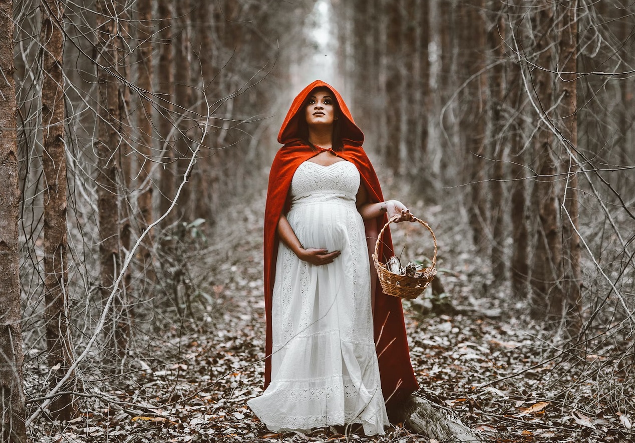 A pregnant woman in a long white dress and bright red cape grips her belly, standing among tall trees.