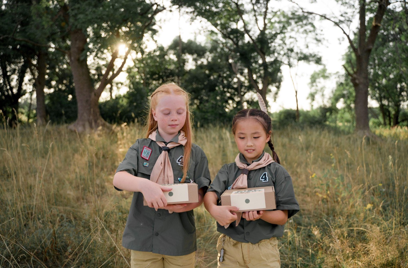 Two Girl Scouts holding boxes in a grassy meadow; image by Cottonbro Productions, via Pexels.com.
