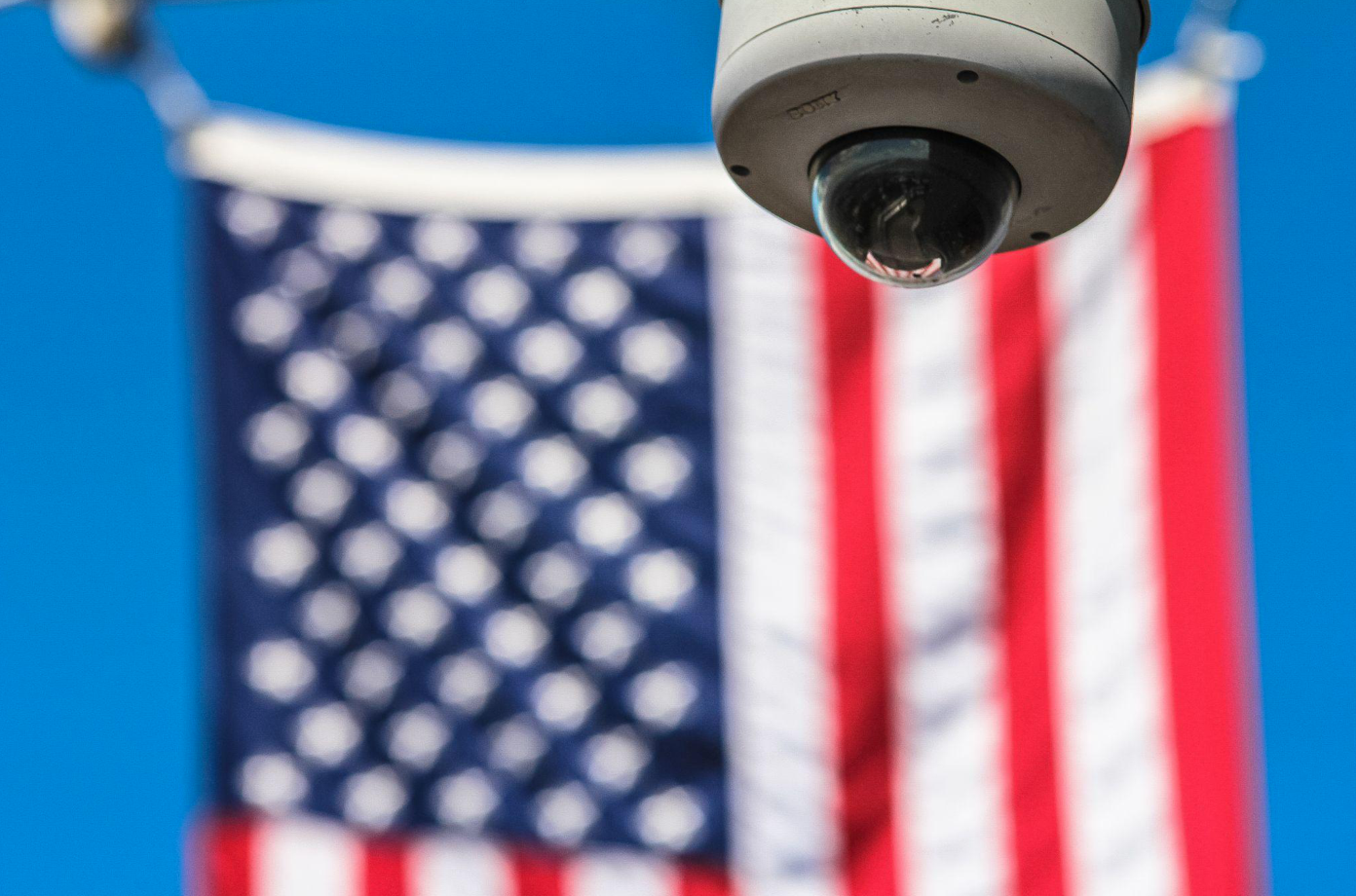 White security camera with blurry American flag as backdrop; image by Francesco Ungaro, via Pexels.com.