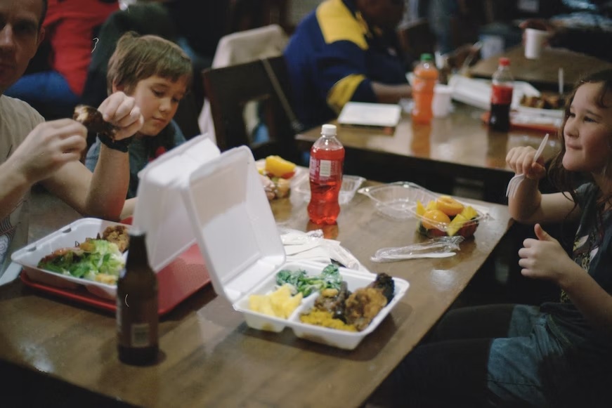 A table full of school age children smiling and eating from lunch containers.