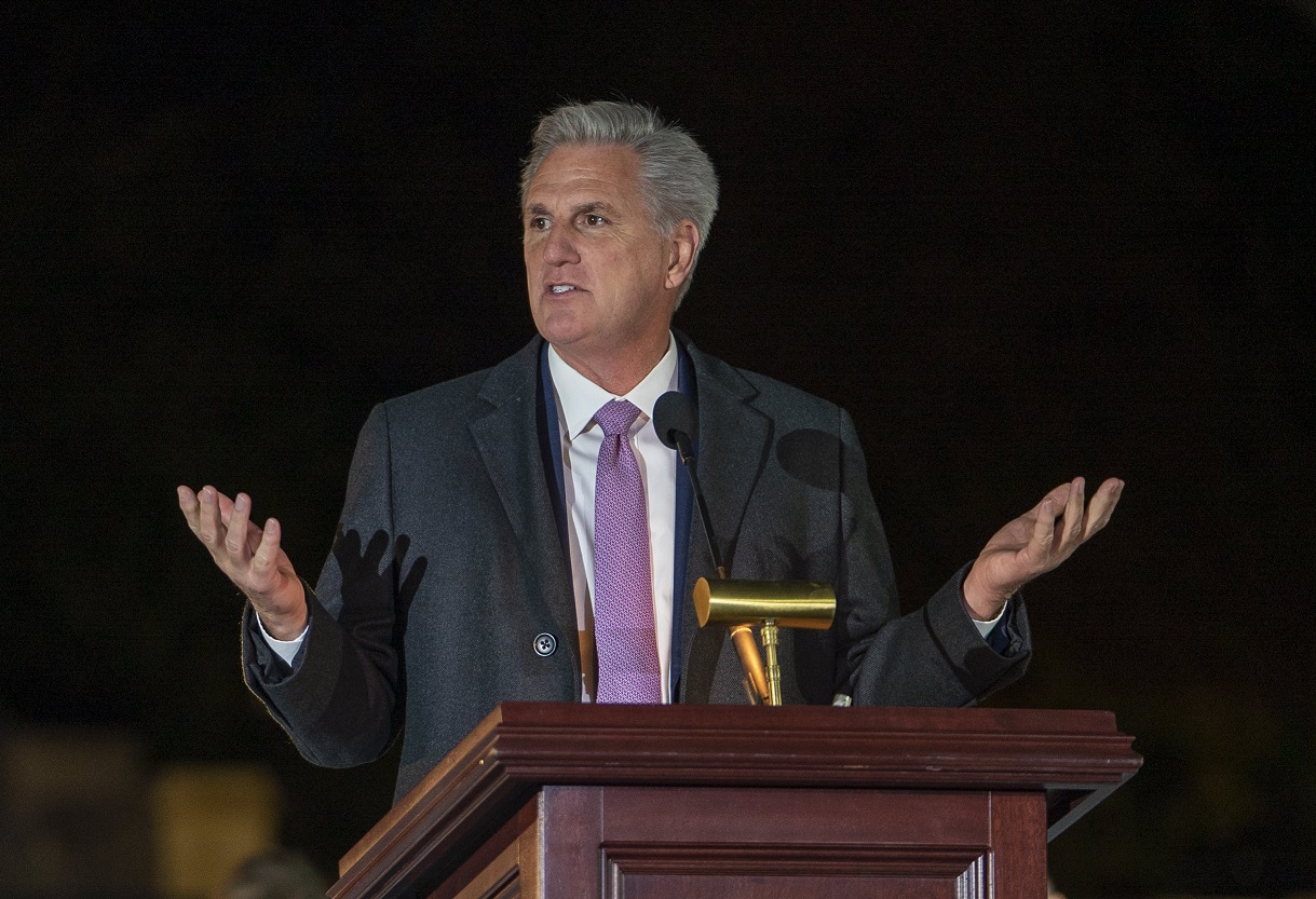Kevin McCarthy wearing a suit and tie, standing behind a podium, with his hands outspread.