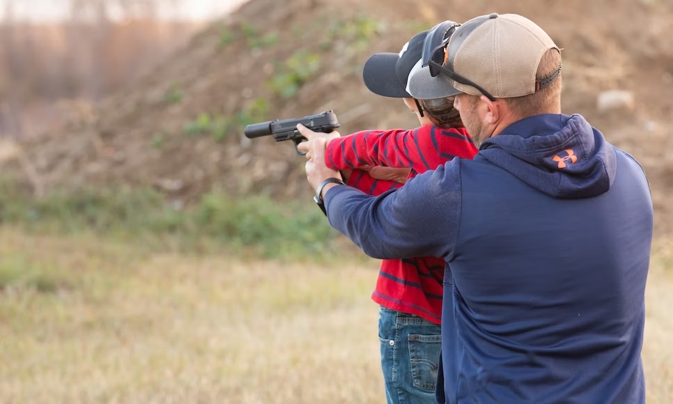 An adult man stands behind a young person in an implied training position, both holding a gun.