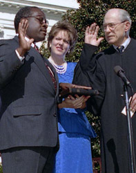 An older white man in a judge's robe swearing in a younger Black man in a suit, accompanied the the background by a white woman in a blue dress.
