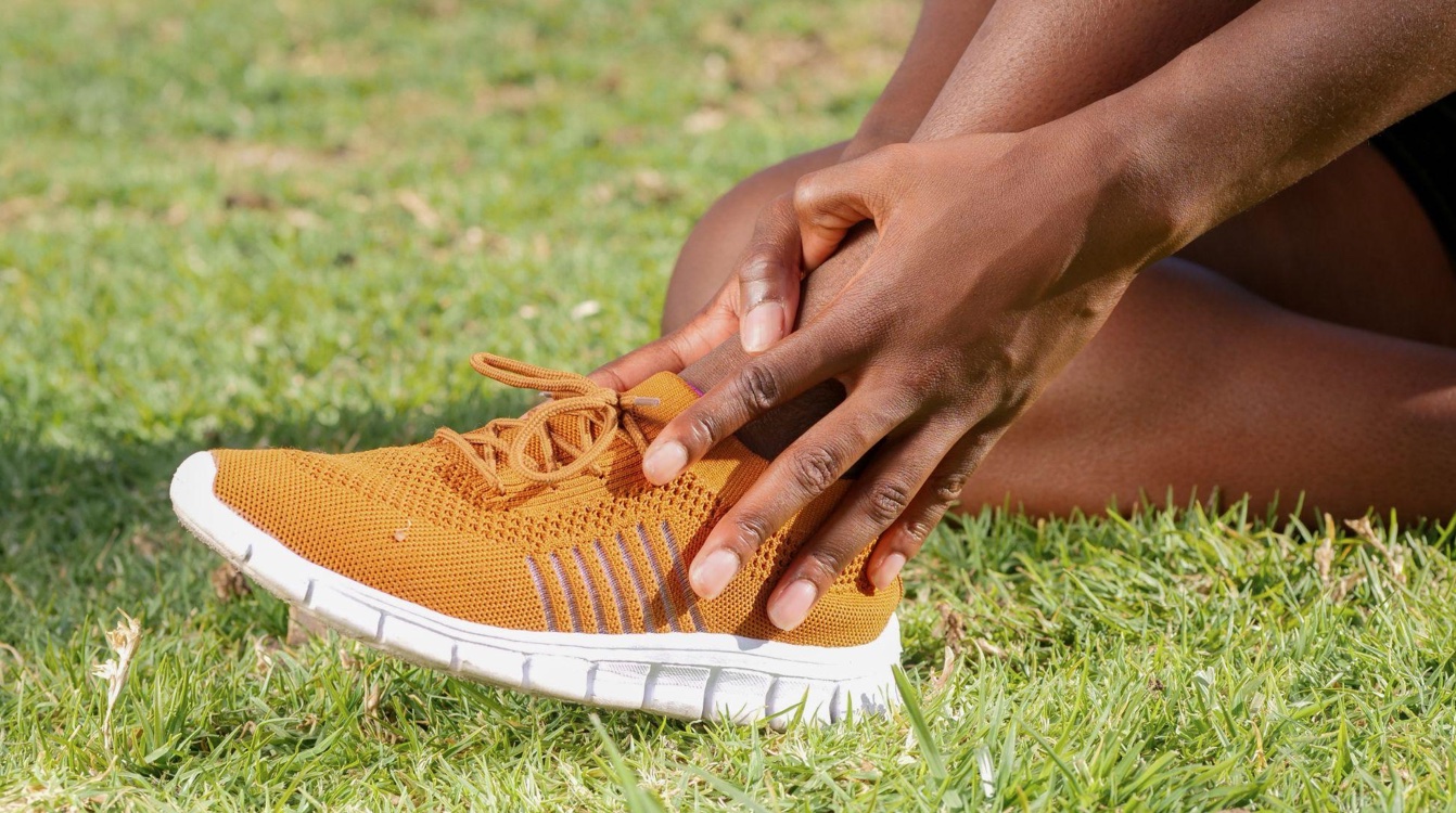 Man in orange sneakers sitting on grass and touching his ankle; image by Kindel Media, via Pexels.com.