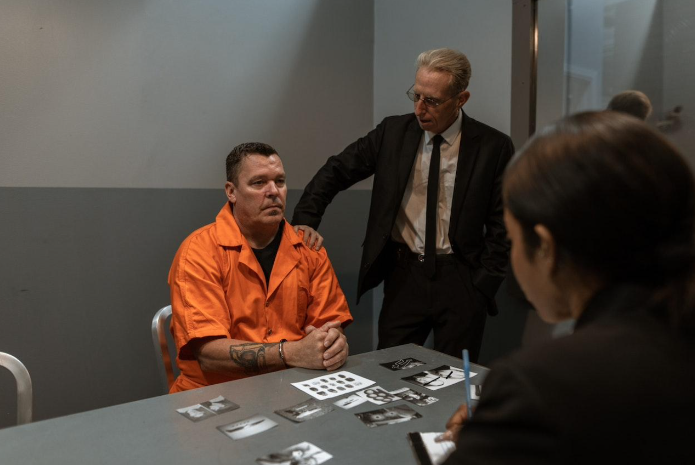 Man in prison orange sitting with man in suit standing and woman sitting across the table; image by RODNAE Productions, via Pexels.com.