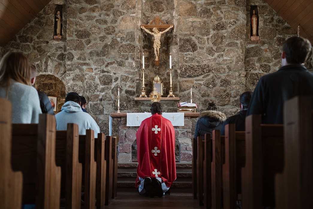 A red-clad figure kneels before an altar in a stone-walled building.