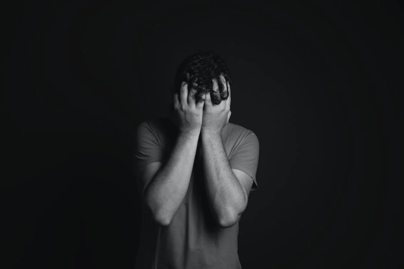 Grey scale photo of man covering his face with hands; image by Daniel Reche, via Pexels.com.