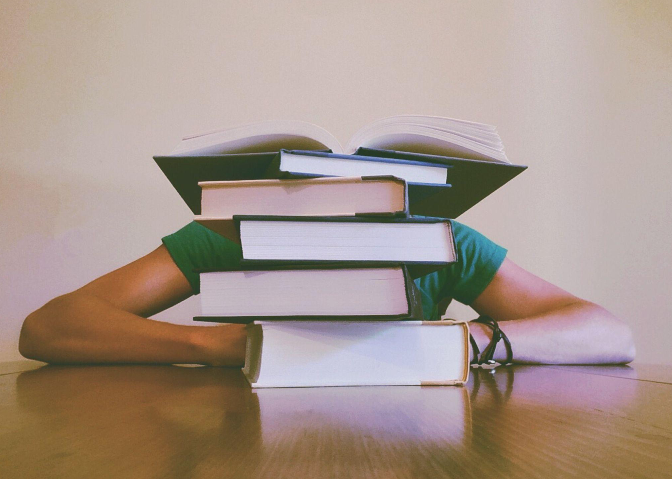Student hiding behind stacks of books; image by Pixabay, via Pexels.com.