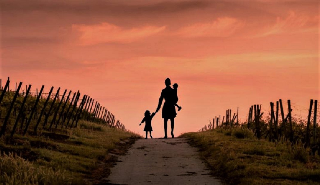 A woman with two children walks along a fence-lined path, silhouetted against an orange sky.