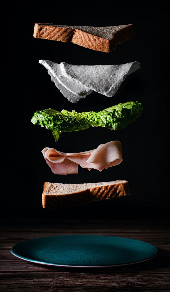 Sandwich ingredients, including bread, lettuce, deli meat, and cheese, falling onto a plate.