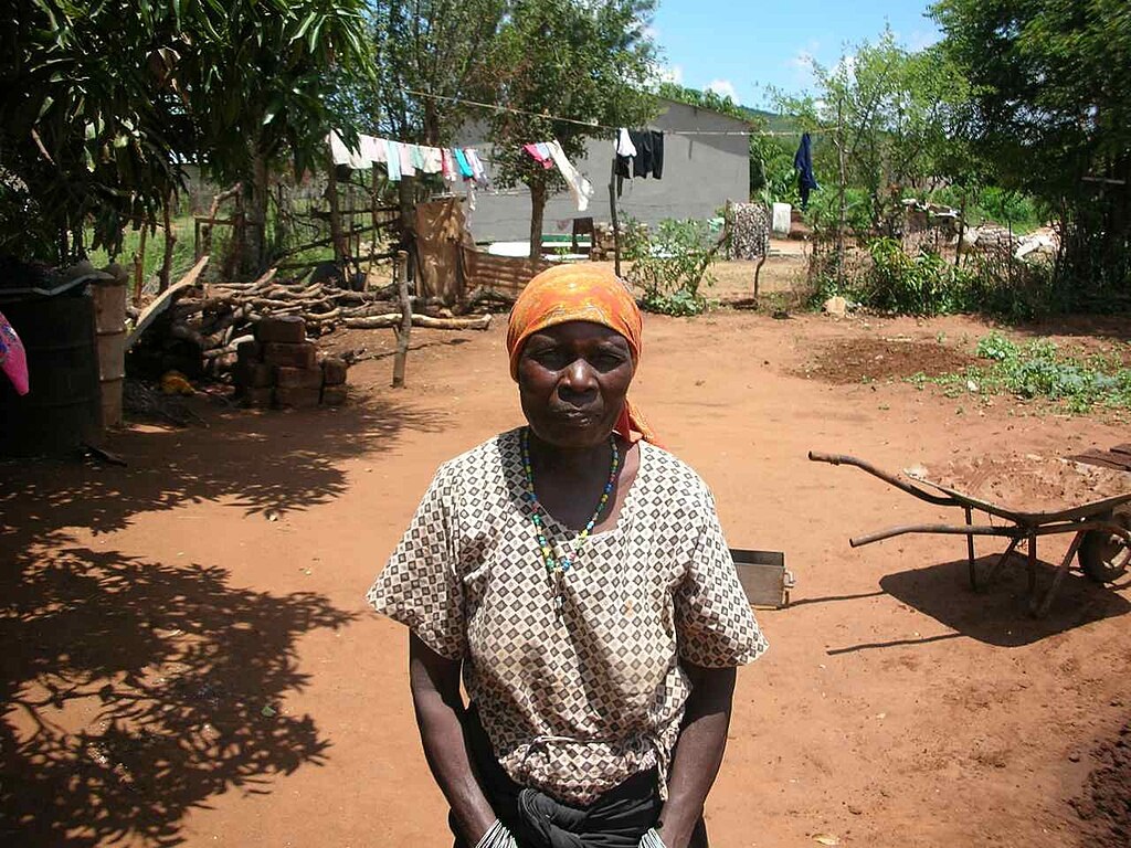A woman with a patterned shirt and a head scarf stands in front of a rural dwelling.