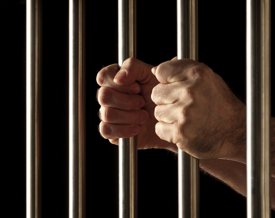 Man's hands gripping jail bars; image by diegoattorney, via Pixabay.com.