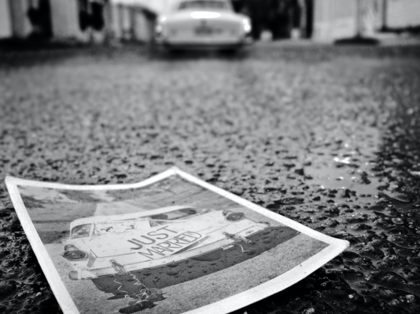 Black and white photo of car driving away, a paper with a picture of the car and a Just Married sign discarded on the pavement; image by Eduardo Sáchez, via Unsplash.com.